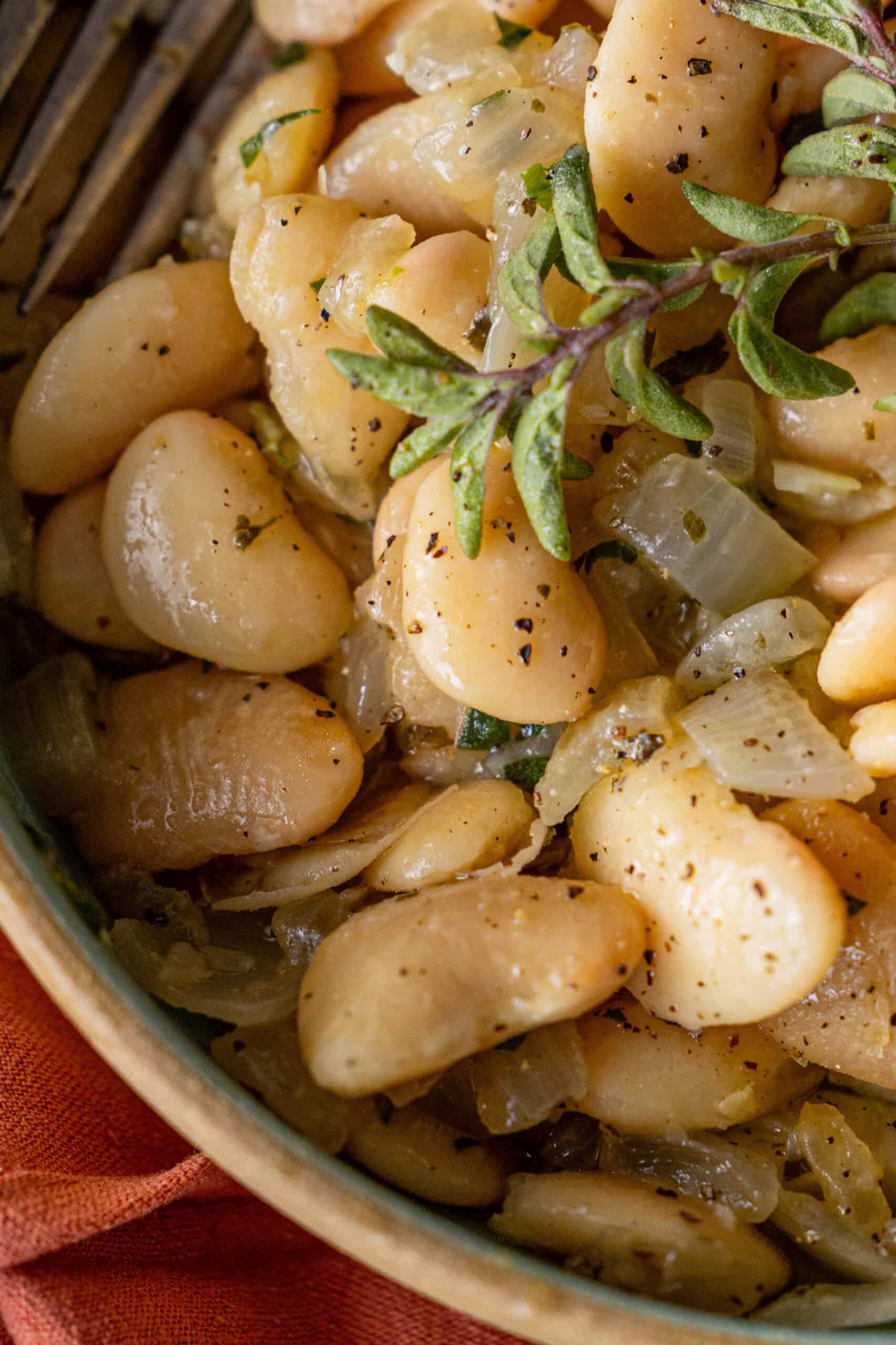 Close-up of cooked butter beans mixed with chopped onions and a hint of garlic, garnished with fresh oregano, in a ceramic bowl.