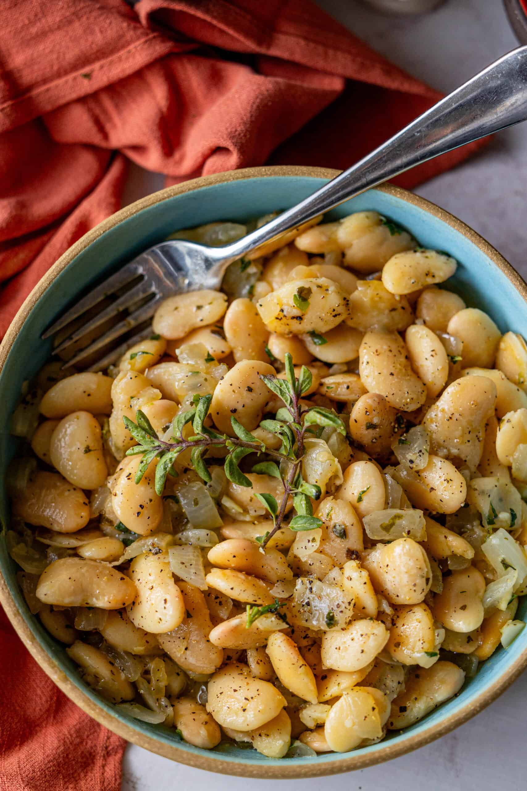 A bowl of cooked butter beans garnished with oregano and herbs, served with a fork on the side. An orange cloth is partially visible in the background.