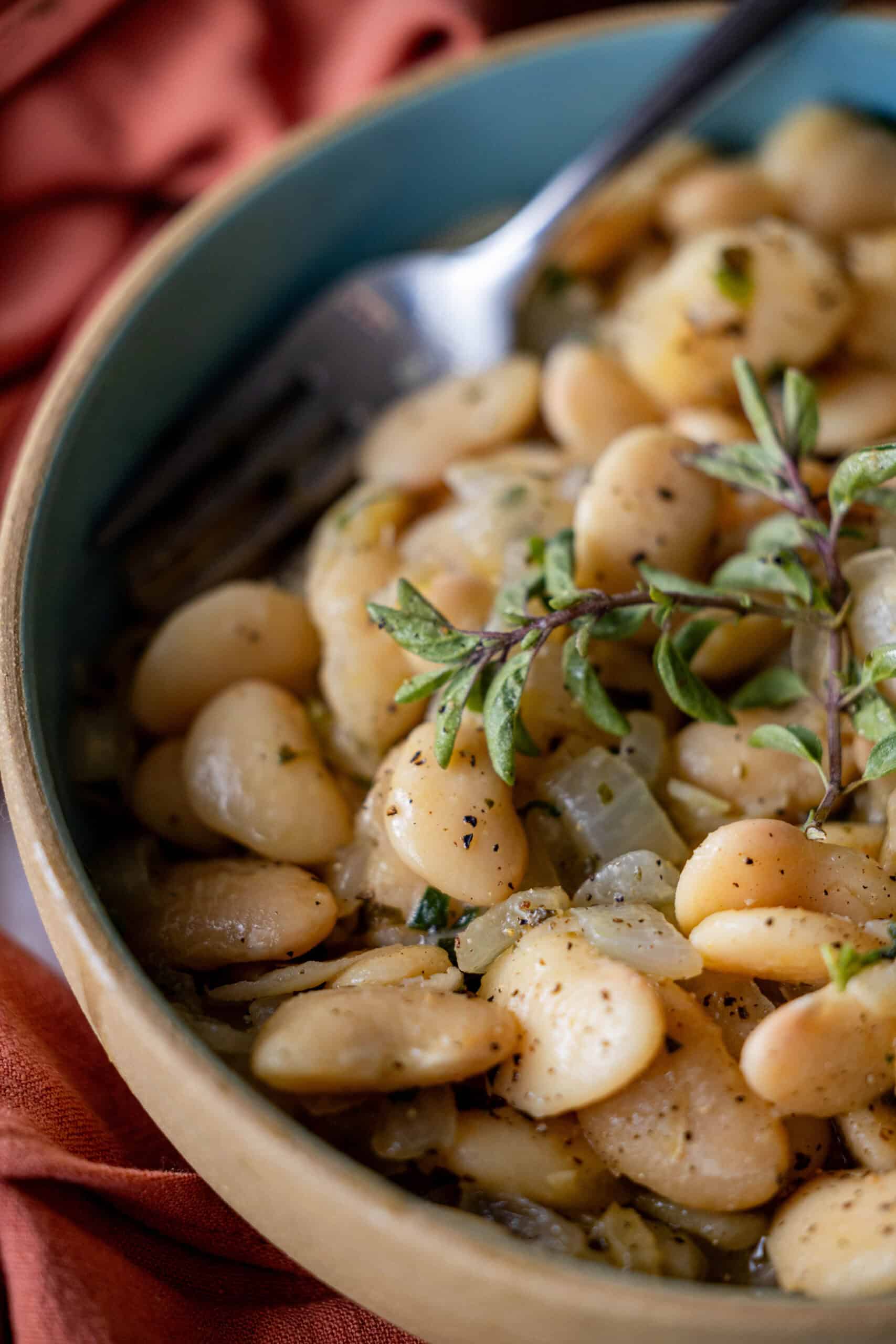 A bowl of seasoned butter beans garnished with fresh herbs and a hint of oregano, with a fork resting inside.