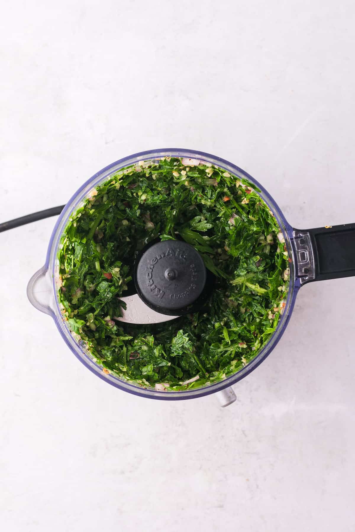 A food processor with chopped greens inside, perfect for whipping up a fresh chimichurri sauce, viewed from above on a light surface.