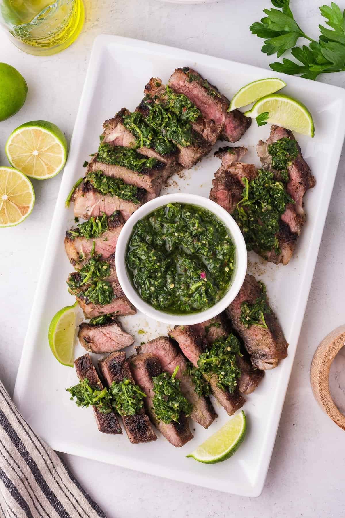 Sliced grilled steak topped with vibrant chimichurri sauce, elegantly arranged on a rectangular plate and garnished with lime wedges. A small bowl of extra chimichurri sits invitingly at the center, adding a burst of flavor to every bite.