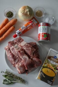 A display of ingredients featuring raw meat, including country style ribs, carrots, onion, tomato paste, canned tomatoes, chicken broth, seasoning, thyme, paprika, and salt on a white surface.