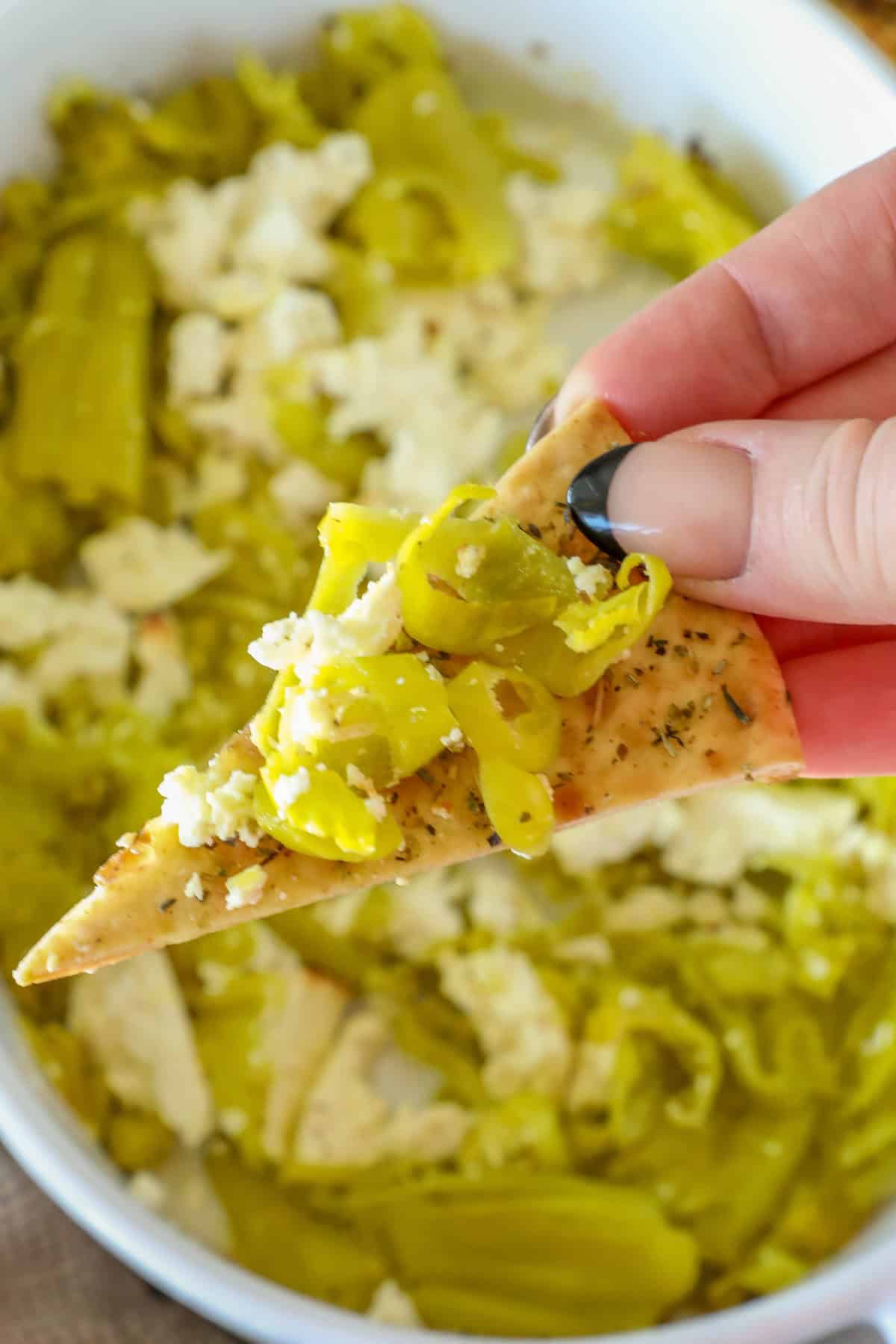 A hand holds a chip topped with sliced green peppers and crumbled cheese over a bowl of pepperoncini baked feta dip.