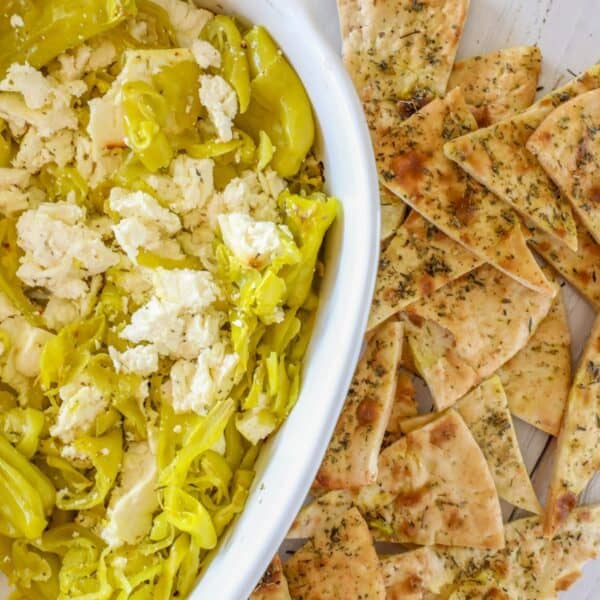 A dish of crumbled feta cheese made into a delightful baked feta dip, accompanied by sliced pepperoncini and pieces of seasoned flatbread.