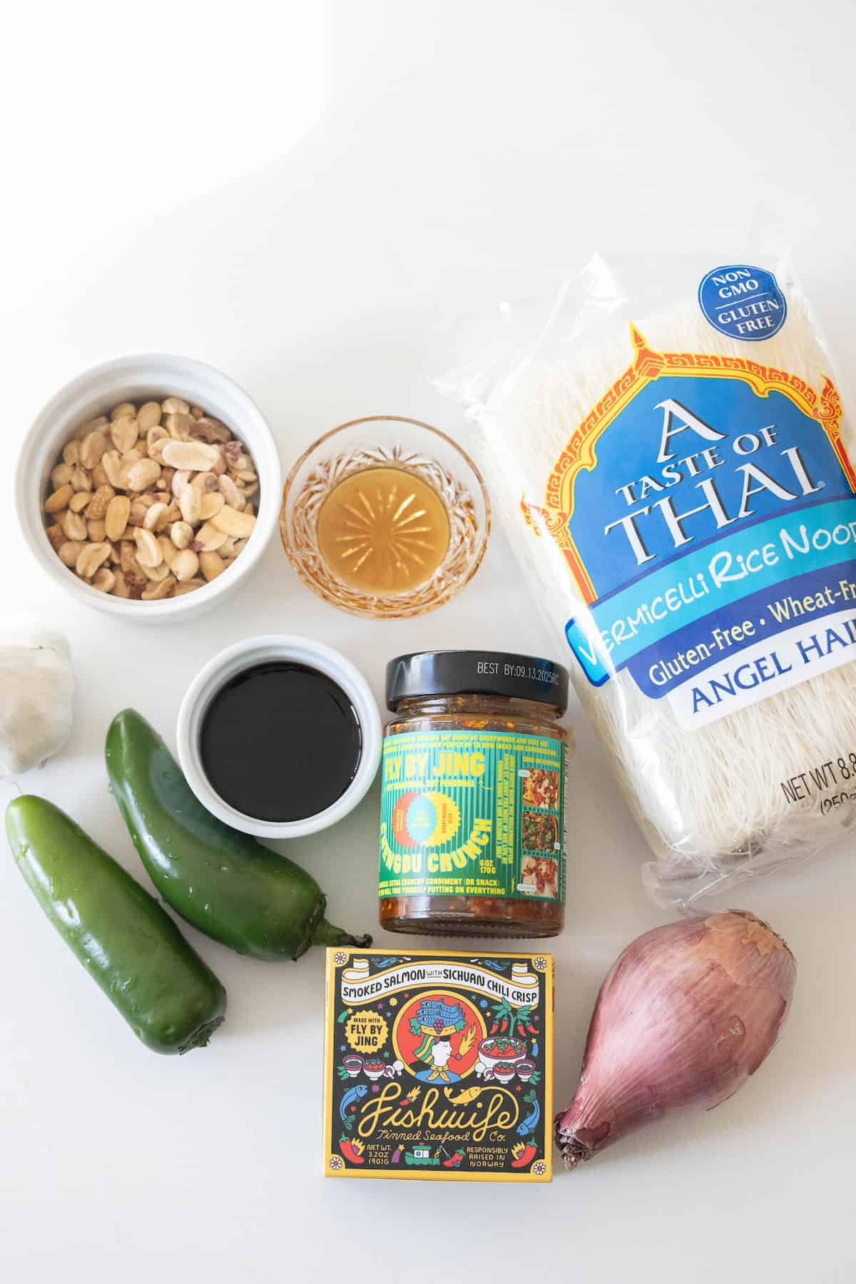 Ingredients for a dish including vermicelli rice noodles, peanuts, jalapeños, garlic, soy sauce, fish sauce, a shallot, and a jar of red curry paste with an added touch of chili arranged on a white surface.