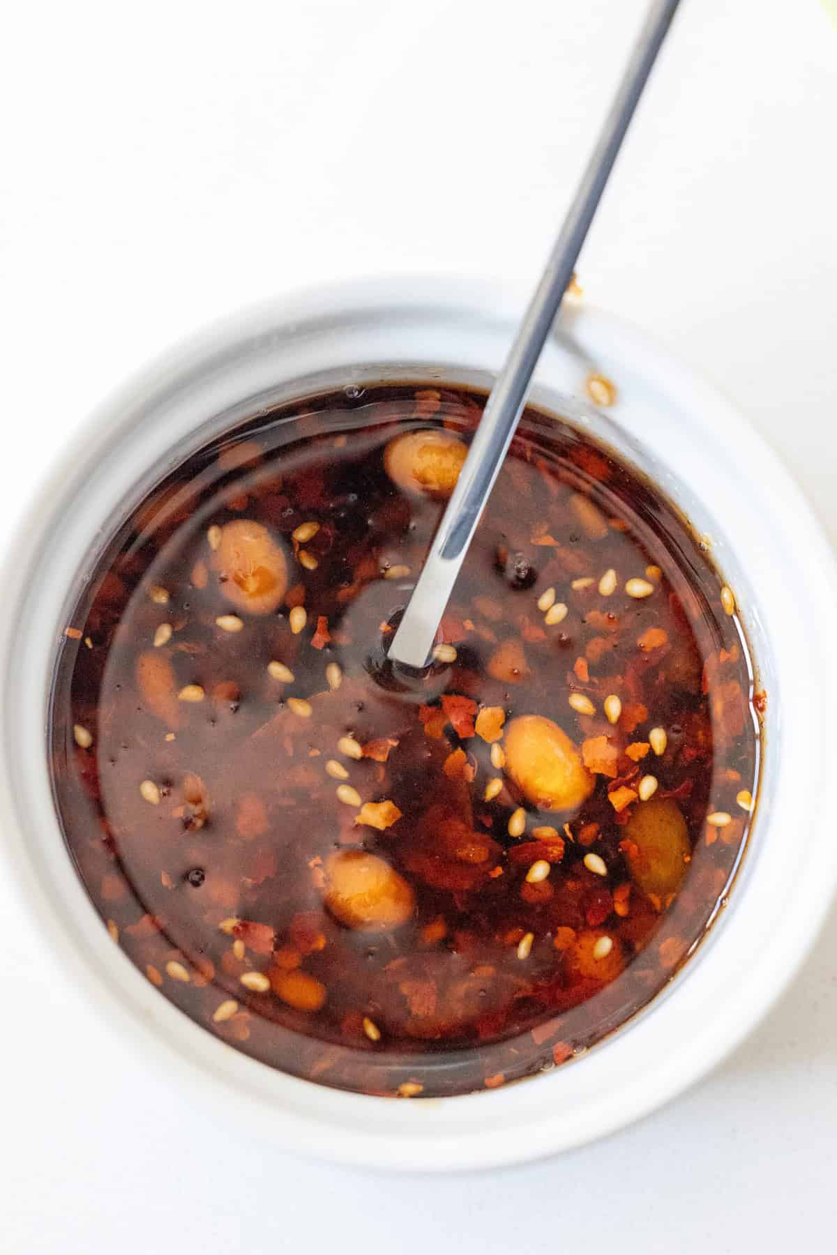 A white bowl filled with chili oil, featuring sesame seeds, chili flakes, and garlic cloves, sits ready to spice up any dish. A spoon is immersed in the vibrant mixture, promising added flavor to your salmon or noodles.