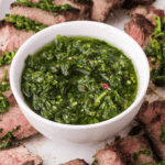 A bowl of vibrant chimichurri sauce, crafted from fresh ingredients, is surrounded by perfectly cooked steak slices on a white plate.