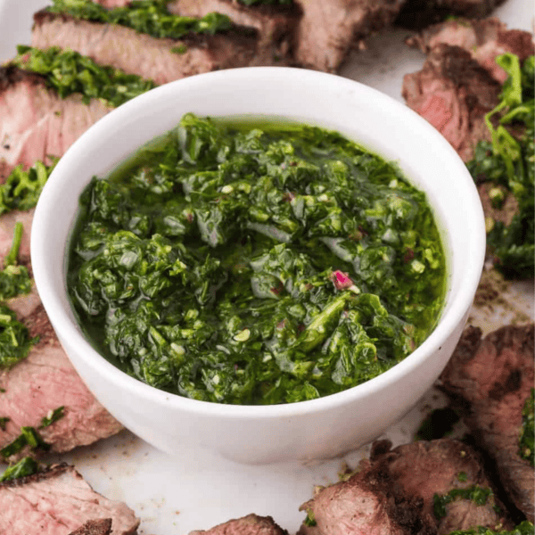 A bowl of vibrant chimichurri sauce, crafted from fresh ingredients, is surrounded by perfectly cooked steak slices on a white plate.