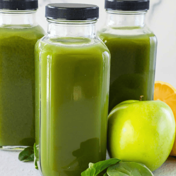 Three bottles of immunity-boosting green juice sit next to a crisp green apple, fresh spinach leaves, and a lemon half on a light surface.