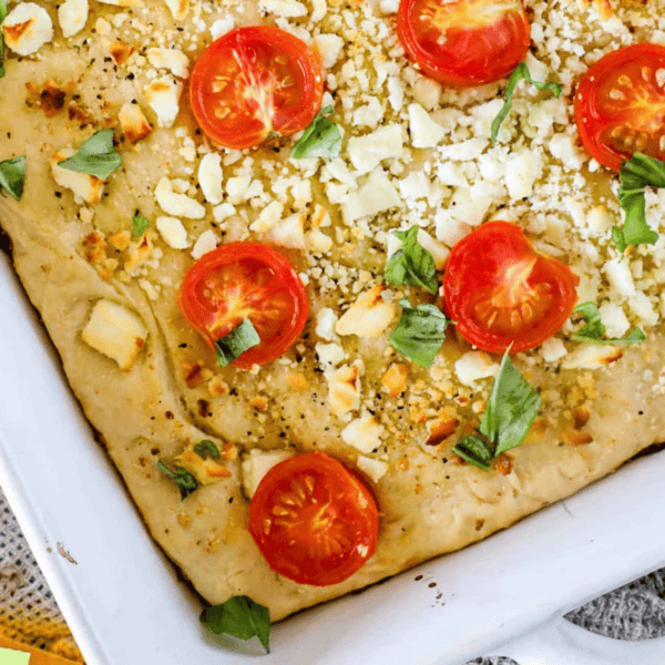 Close-up of a baked Mediterranean focaccia bread topped with sliced tomatoes, crumbled cheese, and fresh basil leaves in a white baking dish.