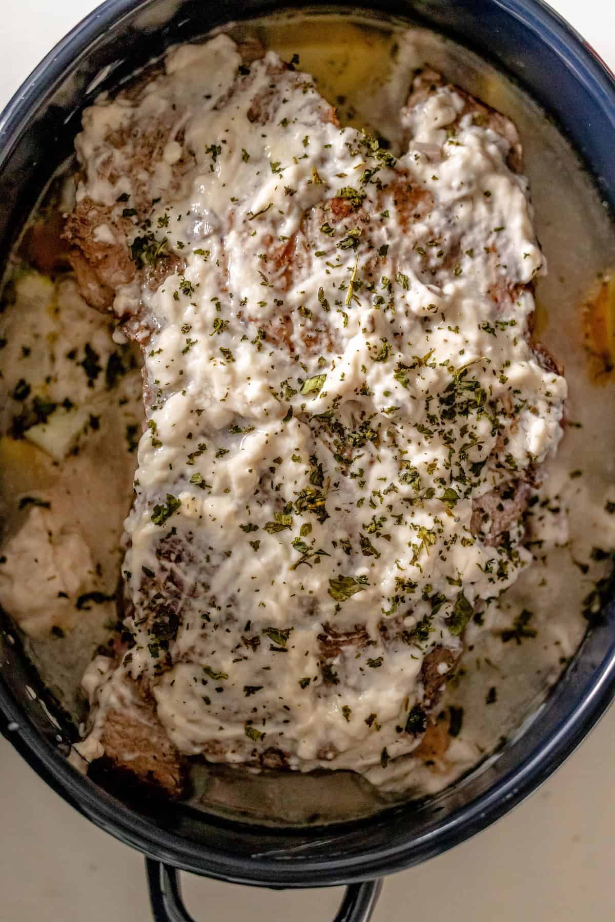 A cooked dish in a pan, featuring a large piece of pot roast topped with creamy white sauce and sprinkled with herbs.