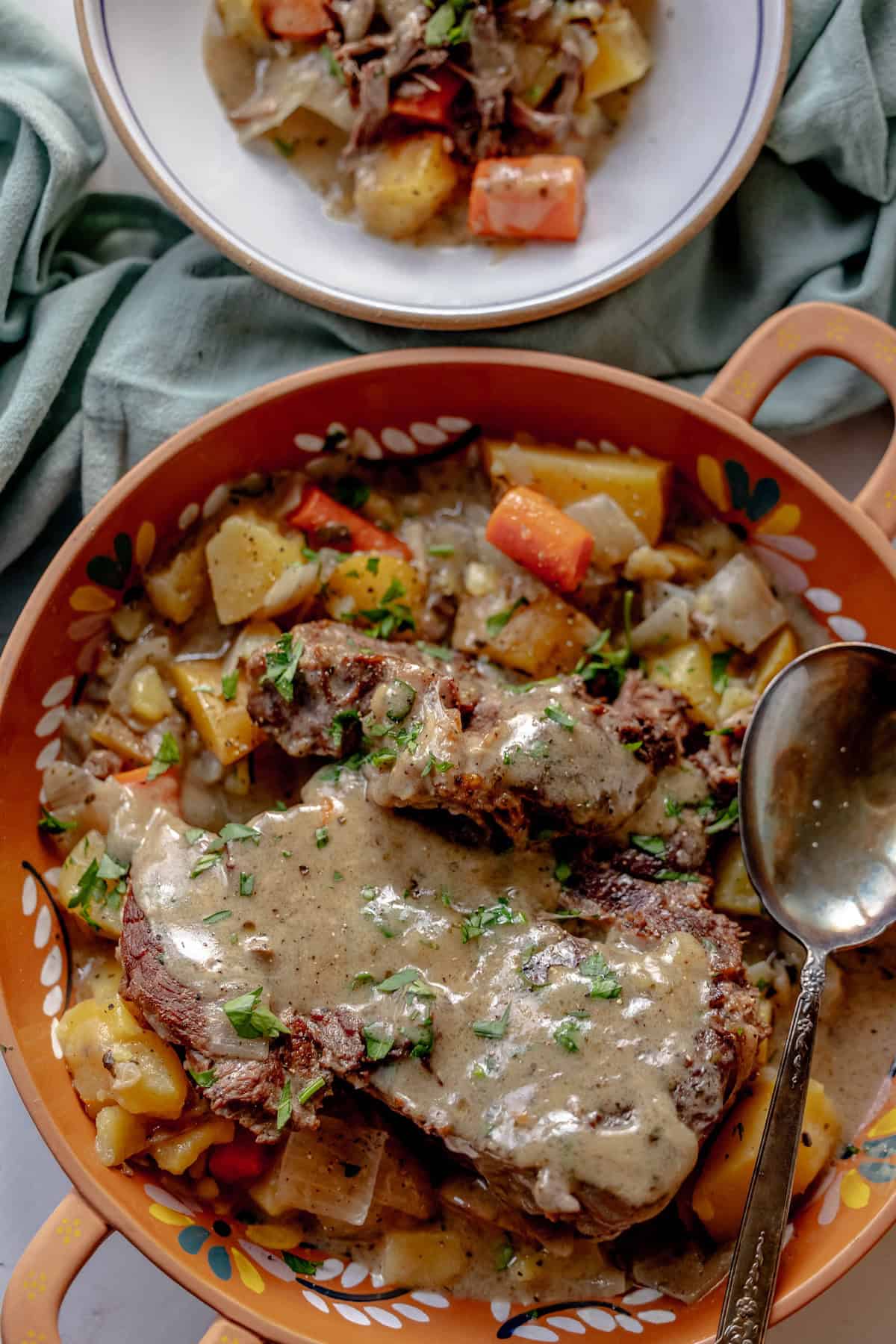 A bowl featuring a savory pot roast dish covered in rich sauce, surrounded by diced potatoes and carrots. A spoon rests on the side. In the background, a plate with a similar dish is partially visible, hinting at the comforting aroma of mushroom soup mingling in the air.