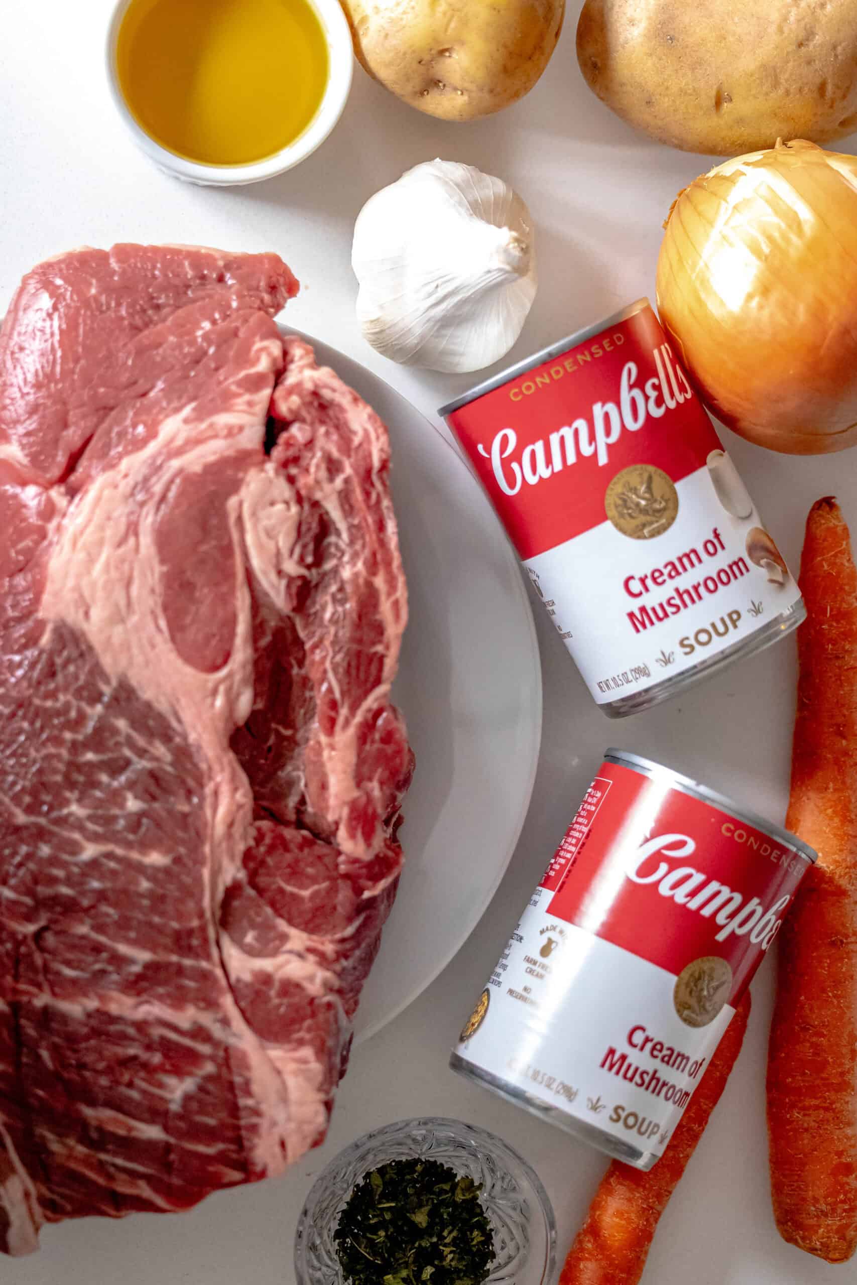 A pot roast ensemble awaits, featuring raw beef, vibrant carrots, aromatic garlic and onion, hearty potatoes, and fresh parsley. Canned mushroom soup completes this culinary canvas on a pristine white surface.
