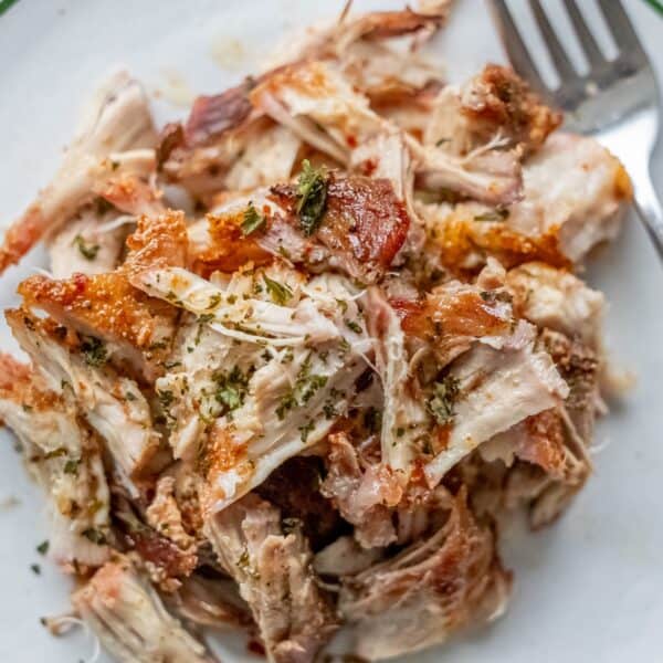 A plate of shredded, oven-roasted chicken garnished with herbs sits elegantly on a white dish, with a fork ready by its side.