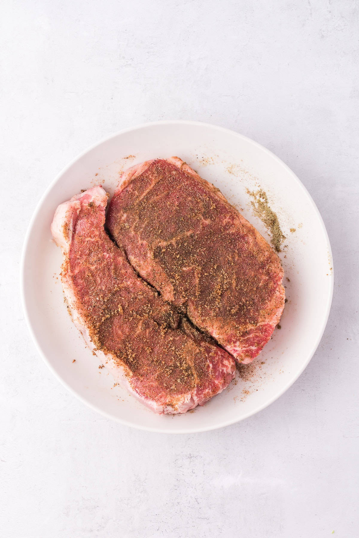 Two seasoned raw steaks on a white plate, ready to meet their fate as succulent grilled steaks, set against a light background.
