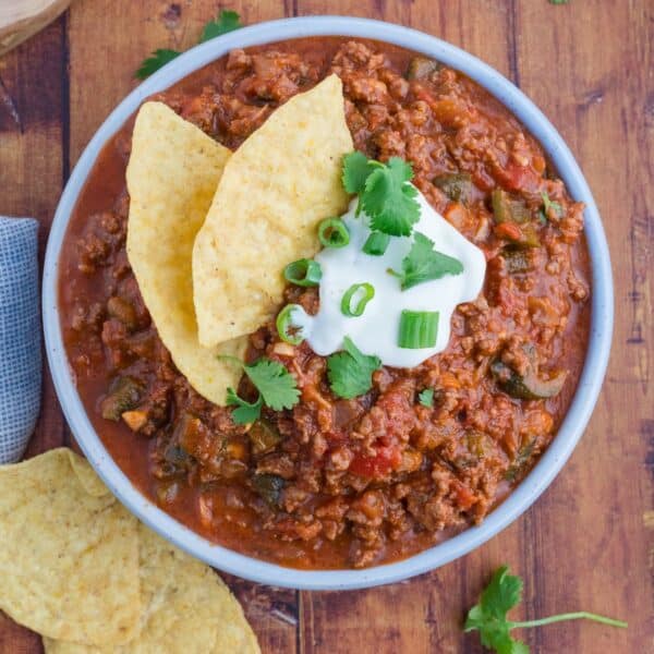 A bowl of chili, spiced with a hint of poblano pepper, is topped with sour cream, chopped green onions, cilantro, and tortilla chips, all beautifully arranged on a wooden surface.