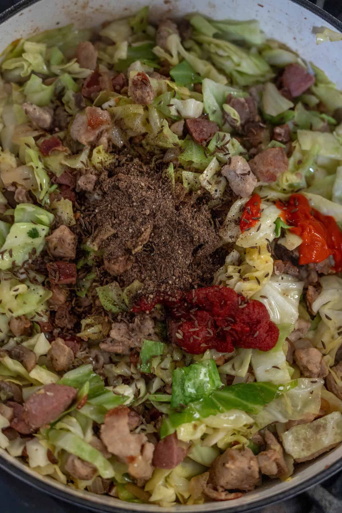 A pot filled with Polish Bigos, featuring chopped cabbage, sausage pieces, ground meat, spices, hot pepper, and tomato paste.