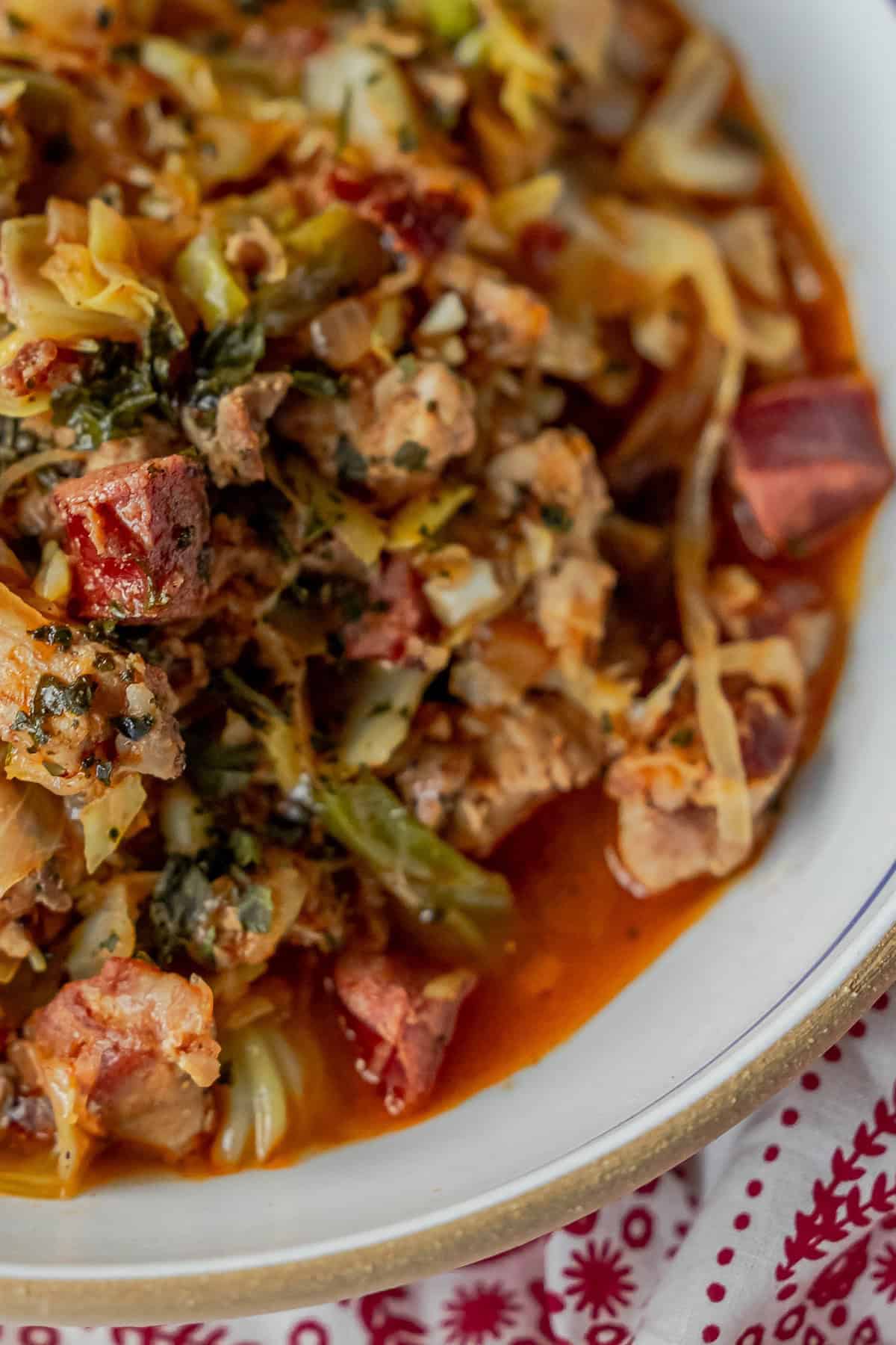 A close-up of a hearty Polish Bigos, also known as Hunters Stew, with chunks of meat, vegetables, and herbs in a thick red sauce. It is served in a white bowl on a red and white patterned tablecloth.