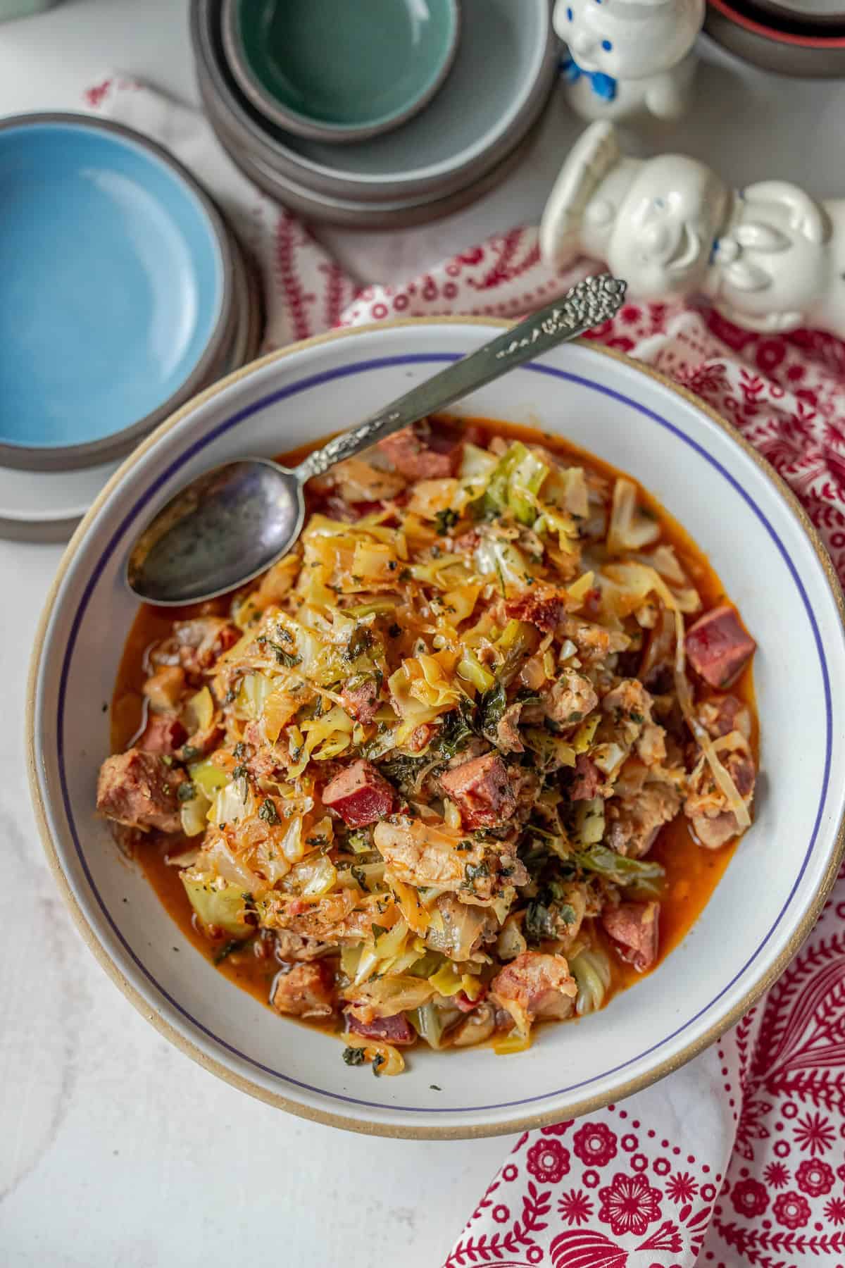 A bowl of hearty hunter's stew, reminiscent of traditional Polish bigos, brimming with vegetables and meat, served with a spoon. Plates and decorative items in the background add a charming touch.
