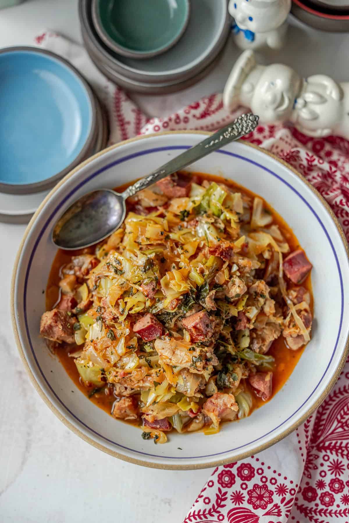 A steaming bowl of Polish Bigos, rich with vegetables and meat, sits invitingly on a red patterned cloth. The hunter's stew is ready to be savored with a spoon, while stacked plates await hungry diners at the table.