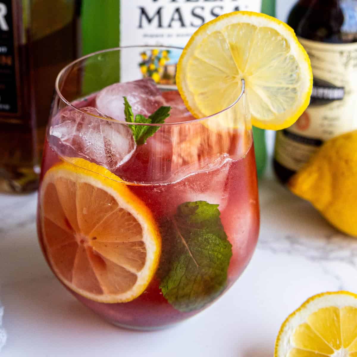 A glass of iced red cocktail garnished with lemon slices and mint leaves. Bottles and a whole lemon are visible in the background.