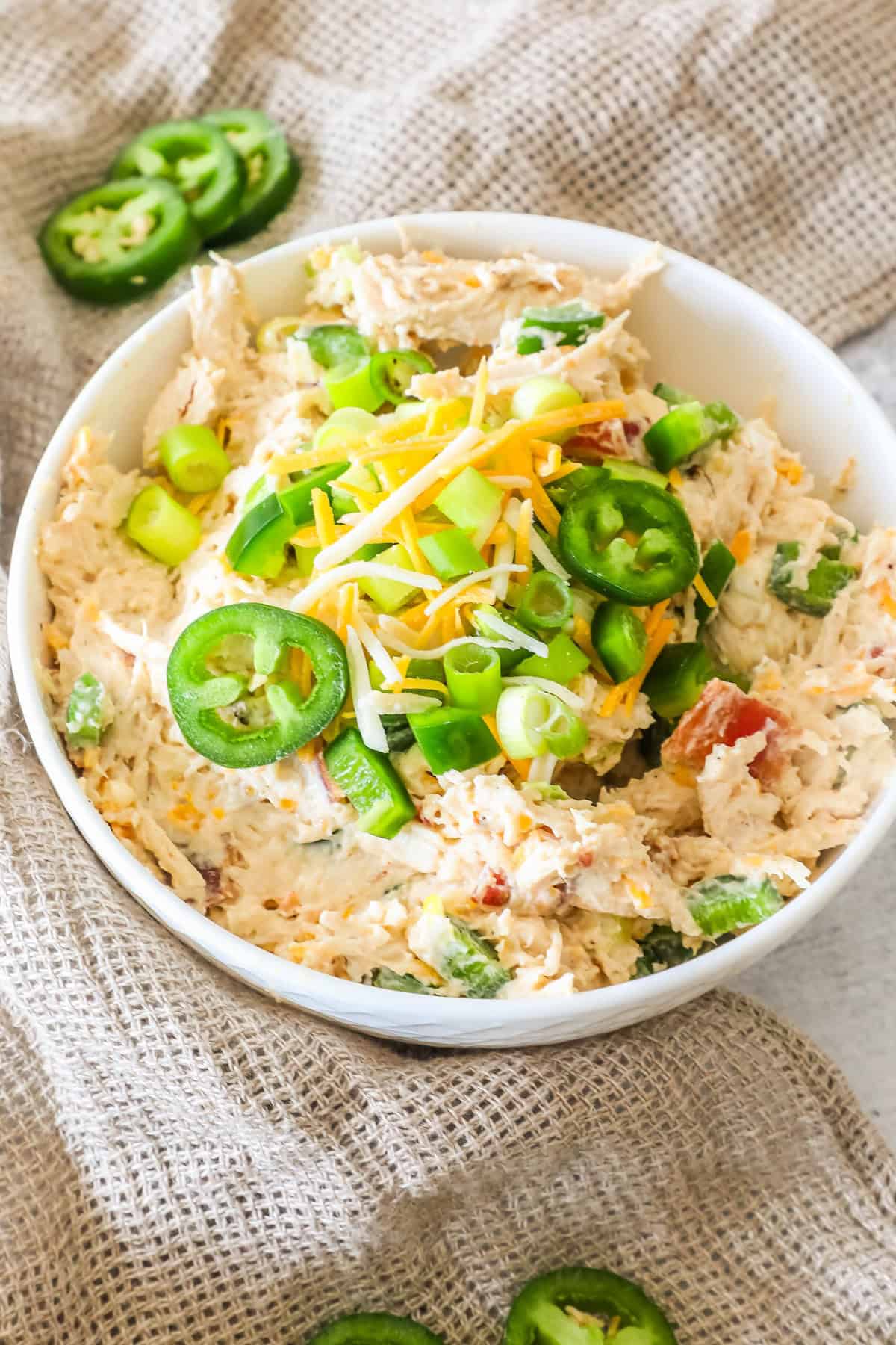 A bowl of chicken salad topped with sliced jalapeños, shredded cheese, and chopped green onions on a burlap cloth.