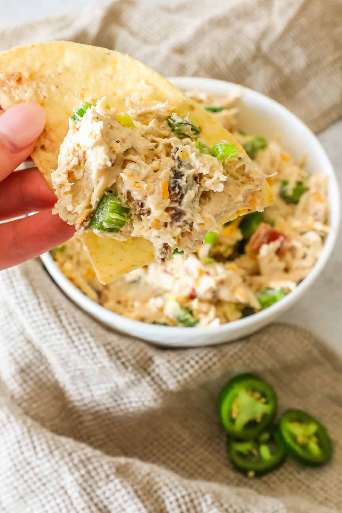 A hand holding a tortilla chip topped with creamy dip over a bowl. Sliced jalapeños and a cloth are in the background.