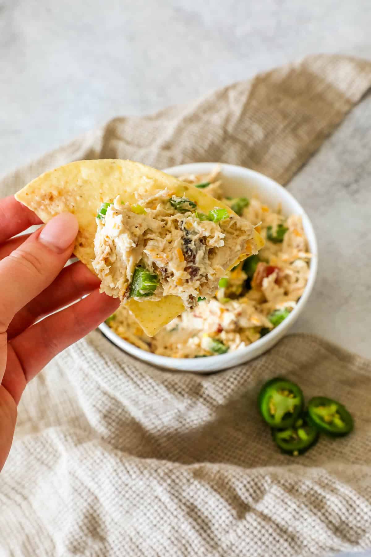 A hand holds a tortilla chip with creamy dip, topped with vegetables, over a dish. Sliced jalapeños are on the side.