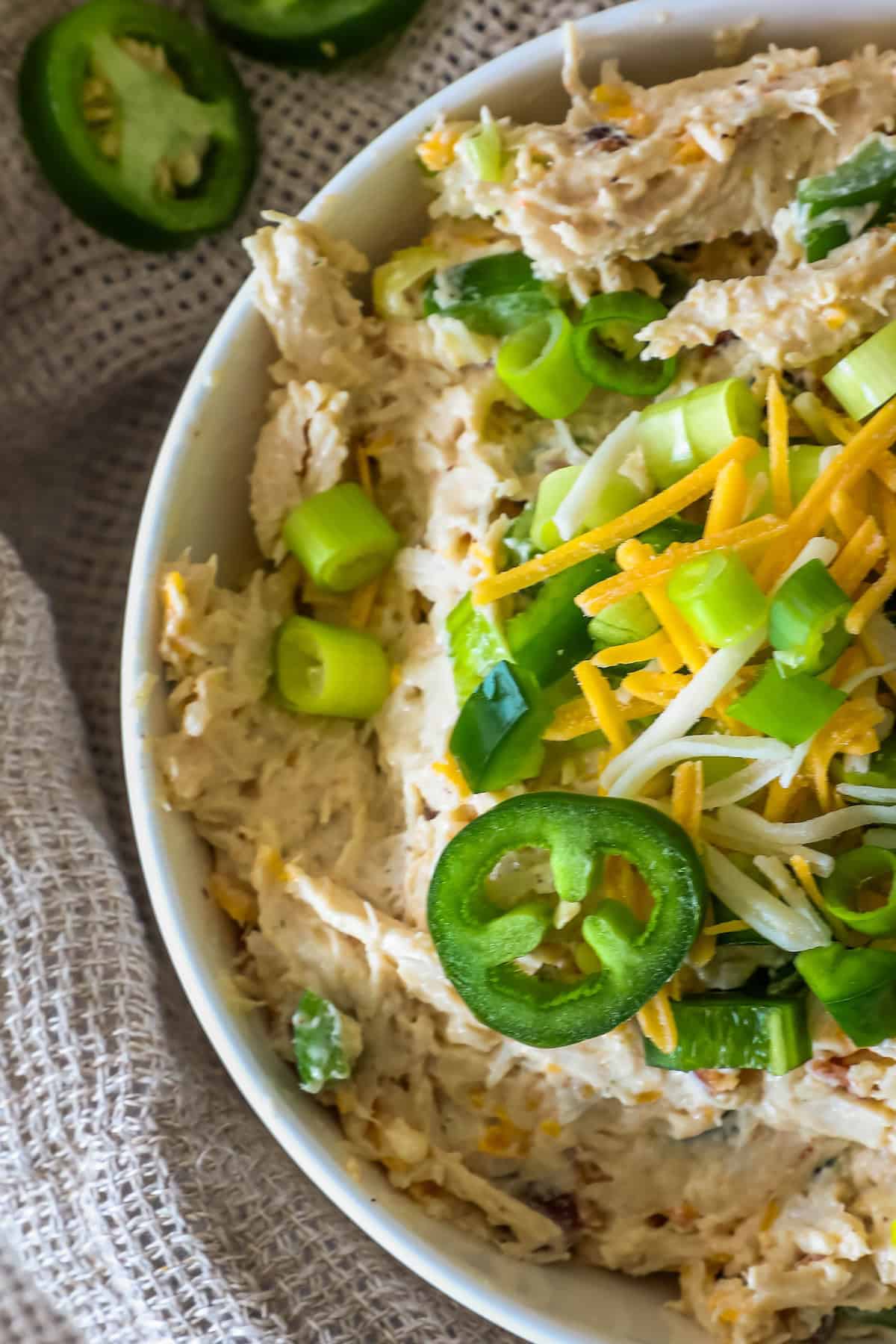 A bowl of creamy dip topped with sliced jalapeños, shredded cheese, and chopped green onions on a textured cloth background.