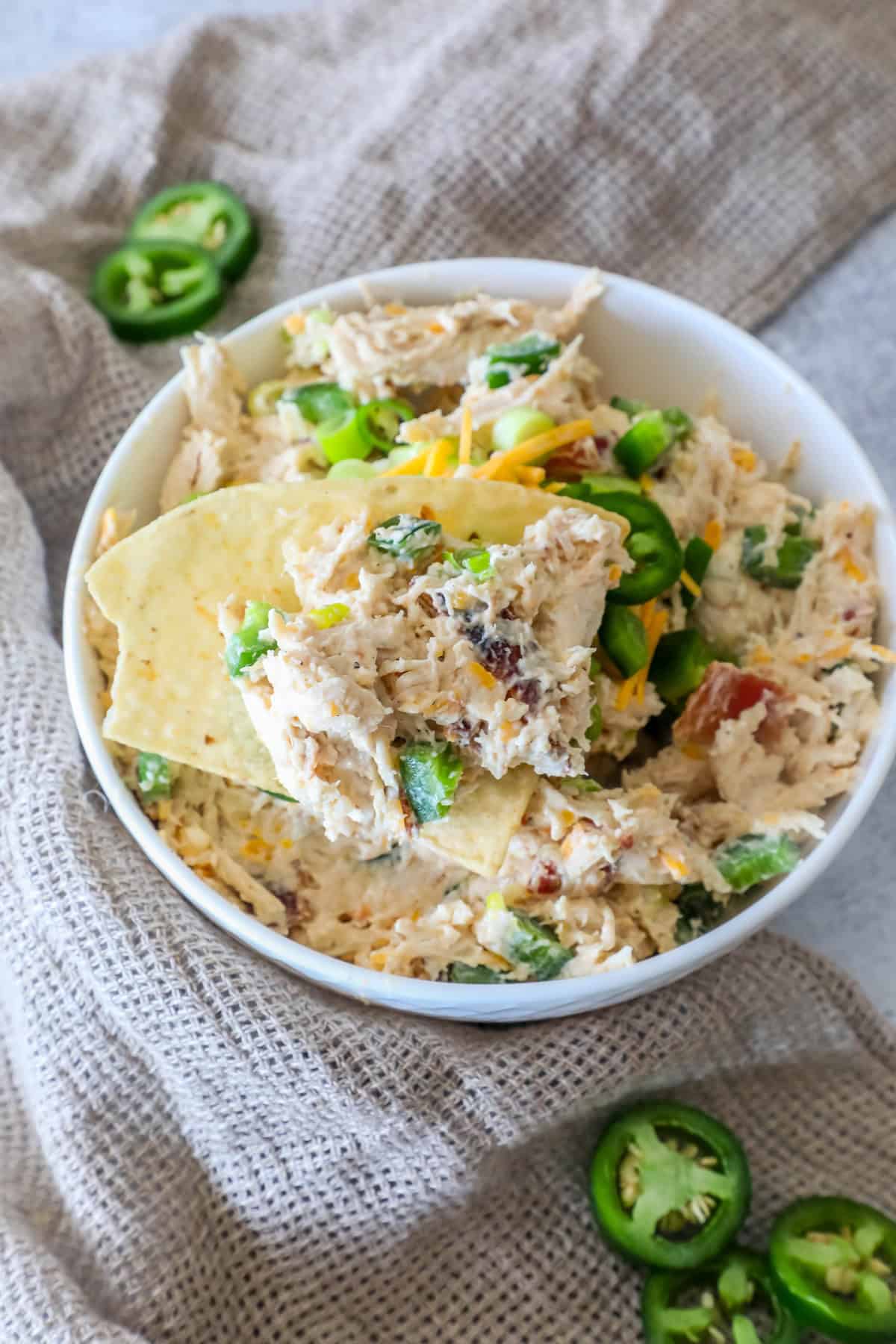 Bowl of creamy chicken dip with sliced jalapeños and shredded cheese, garnished with tortilla chips, set on a textured cloth.