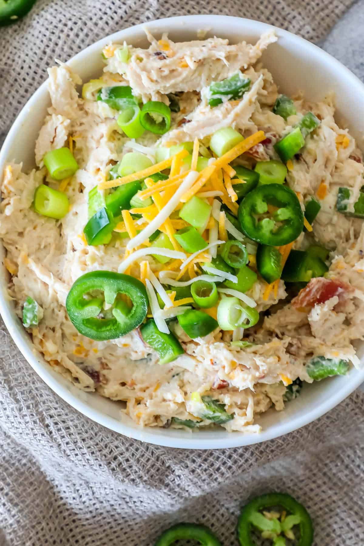 A bowl of chicken dip topped with sliced jalapeños, green onions, and shredded cheese, placed on a burlap surface.