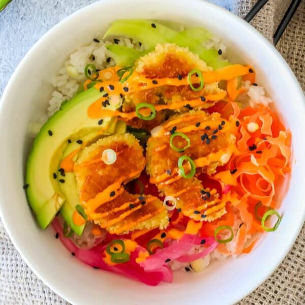 A bowl of rice topped with sliced avocado, fried shrimp with sauce, pickled ginger, shredded vegetables, and black sesame seeds.