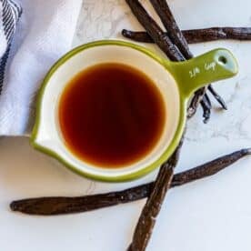 A green measuring cup filled with a dark liquid, labeled 1/4 cup, is placed on a marble surface surrounded by vanilla beans and a striped cloth.