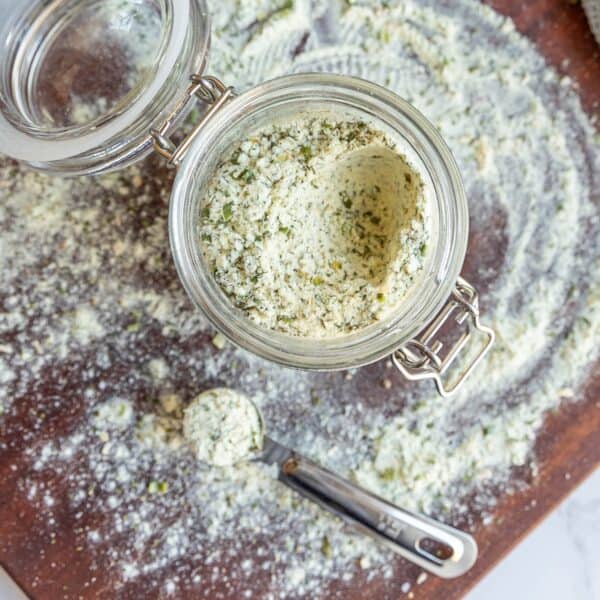 Glass jar of herb-infused salt on a wooden surface with spilled salt and a measuring spoon.