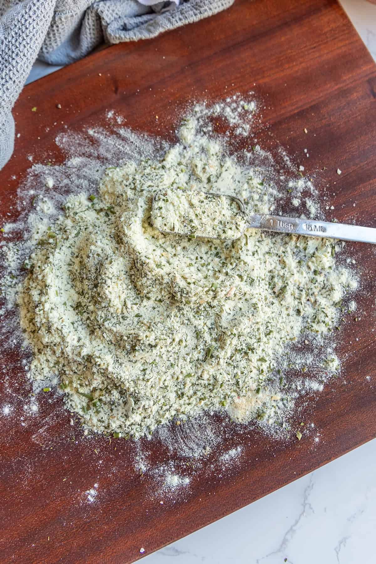 A mix of herbs and flour with a teaspoon on a wooden surface. A gray cloth is partially visible in the upper left corner.