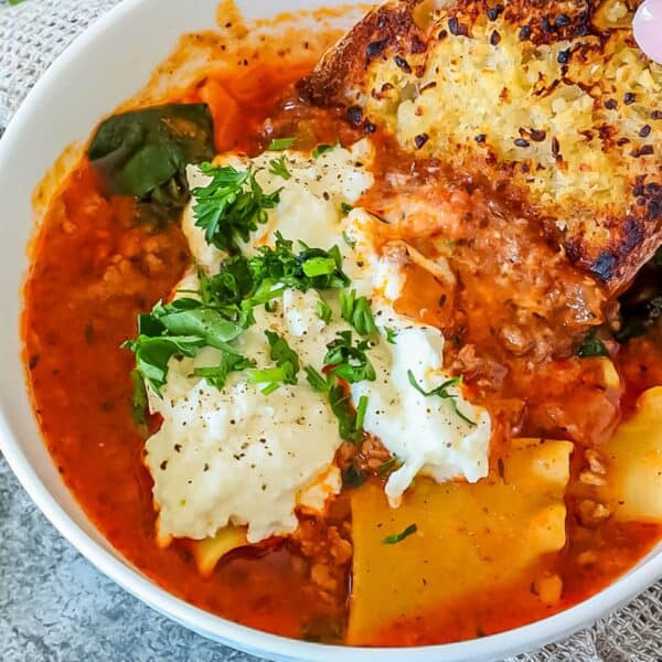 A bowl of lasagna soup topped with ricotta cheese and parsley, accompanied by a slice of garlic bread.