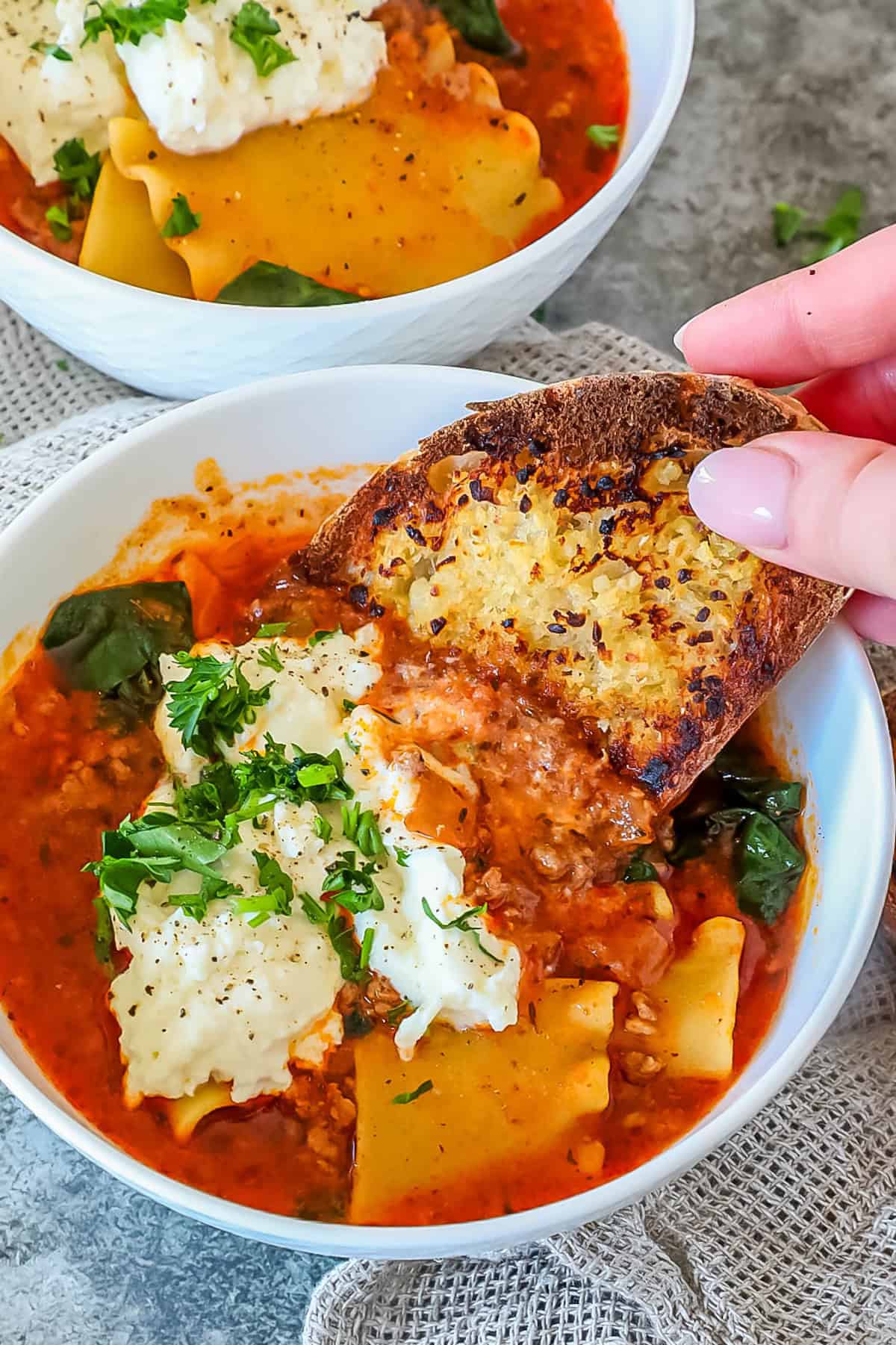 Hand holding toasted bread over a bowl of lasagna soup topped with cheese and parsley. Another bowl with similar content is in the background on a textured surface.