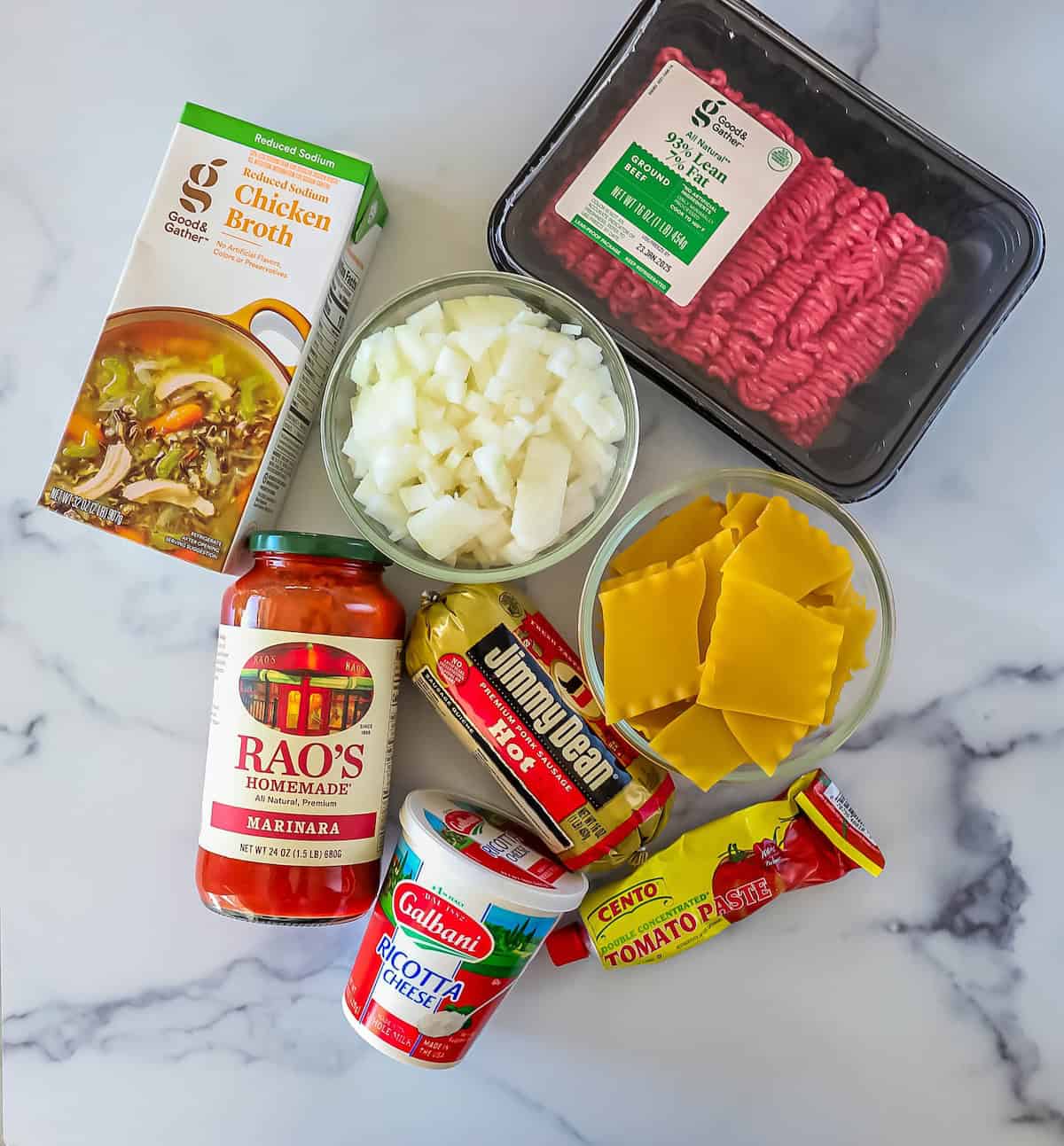 Ingredients on a marble surface: chicken broth, ground beef, chopped onions, lasagna noodles, marinara sauce, ricotta cheese, tomato paste, and a sausage pack.