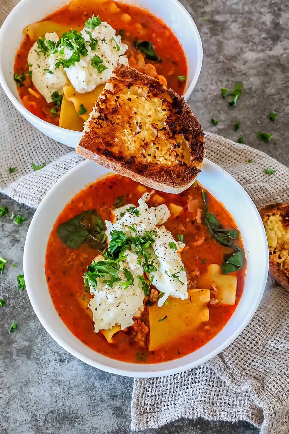 Two bowls of soup with pasta, spinach, and cheese, garnished with herbs. A slice of toasted bread is placed on each bowl.