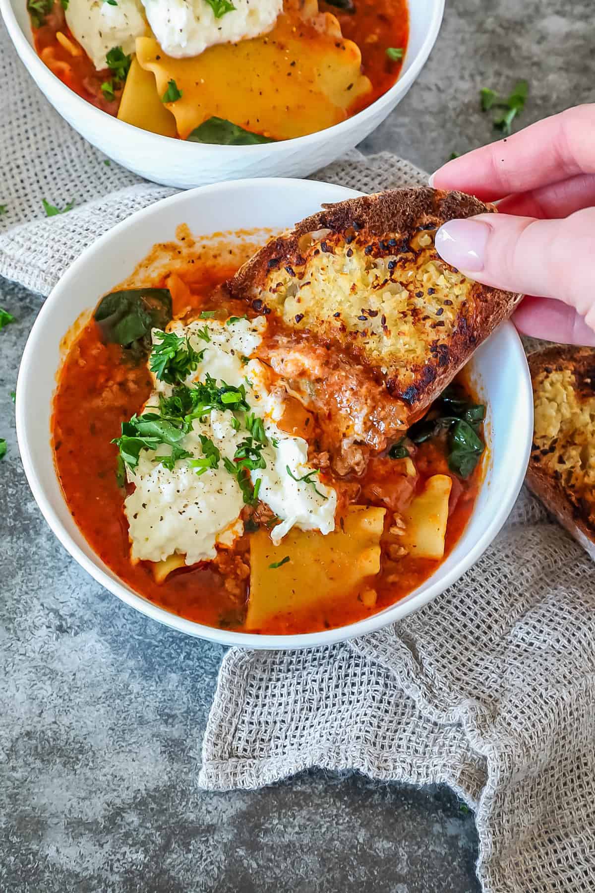 A hand dipping toasted bread into a bowl of lasagna soup topped with ricotta and parsley, on a textured gray surface with another similar bowl in the background.