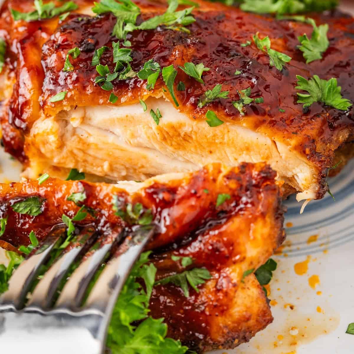 Close-up of a fork cutting into glazed BBQ chicken, garnished with chopped parsley, on a patterned plate.