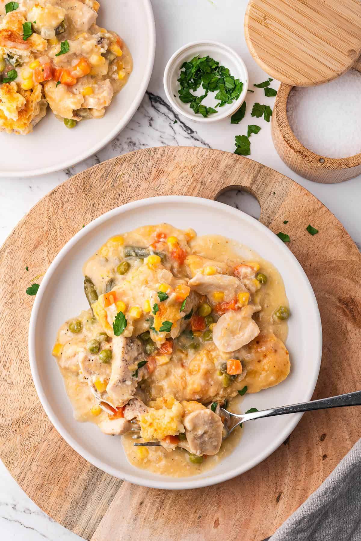 A plate of creamy chicken pot pie casserole with vegetables rests on a wooden board, garnished with parsley, beside a small bowl of herbs and an open salt container.