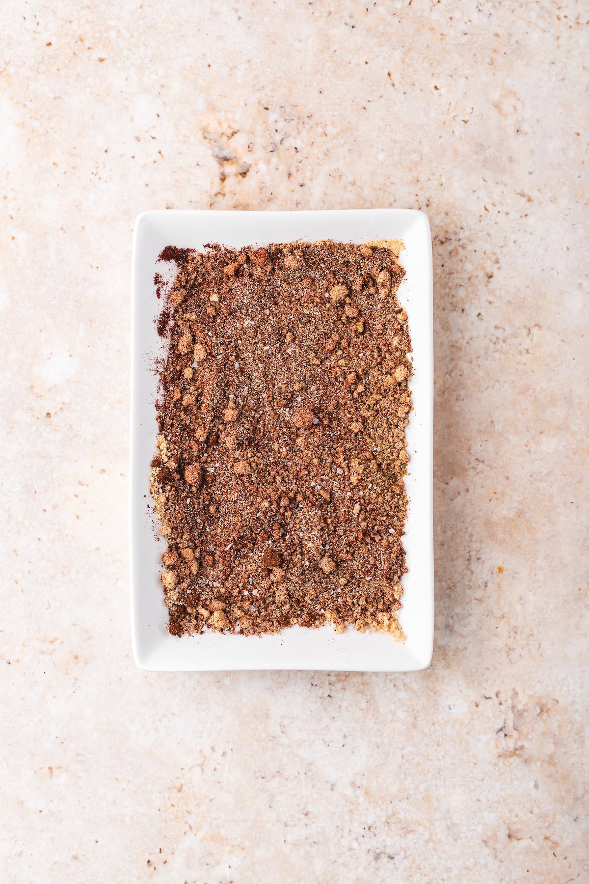 A rectangular white dish filled with ground spices sits on a speckled beige surface, perfect for seasoning your favorite pulled pork recipe in a Dutch oven.