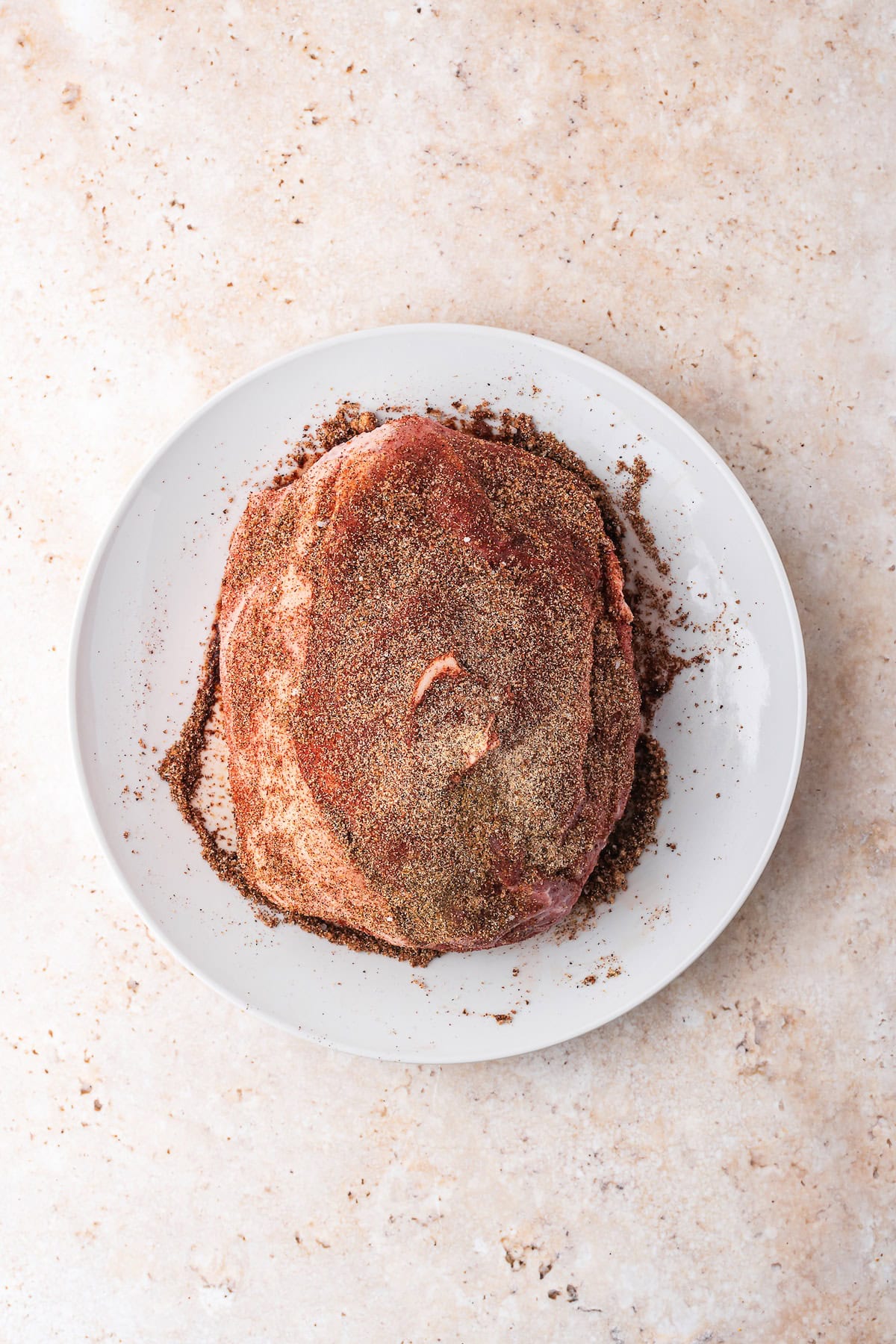 Seasoned raw beef, perfect for your Dutch oven, sits on a white plate against a textured beige surface, ready to transform into a flavorful delight.