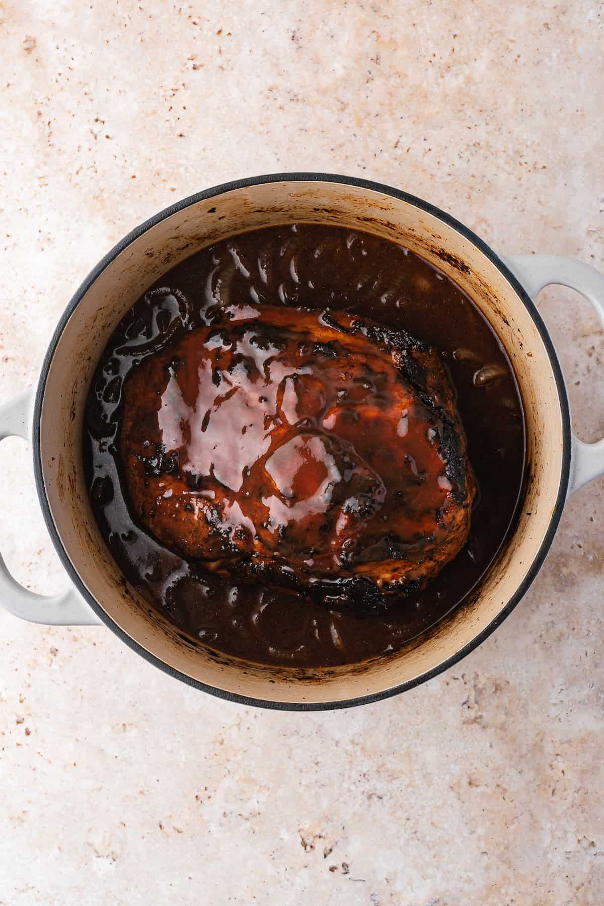 A succulent piece of meat, resembling pulled pork, is bathed in a rich brown sauce inside a round Dutch oven, resting on a light-colored textured surface.