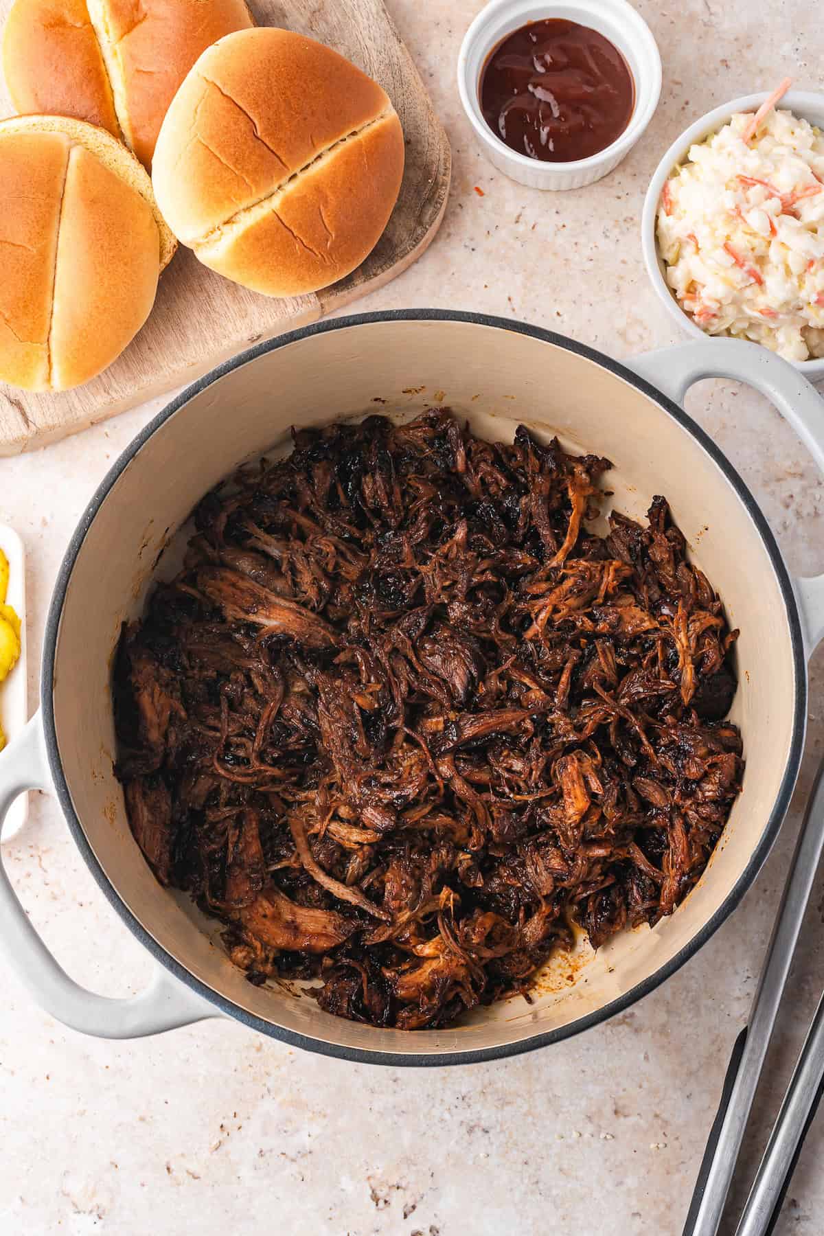 A Dutch oven brimming with tender pulled pork sits beside a wooden board adorned with buns, a dish of coleslaw, and a bowl of barbecue sauce, perfect for crafting that classic recipe everyone loves.