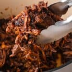 Tongs lift tender, juicy shredded barbacoa beef from a Dutch oven, revealing its rich color and texture.