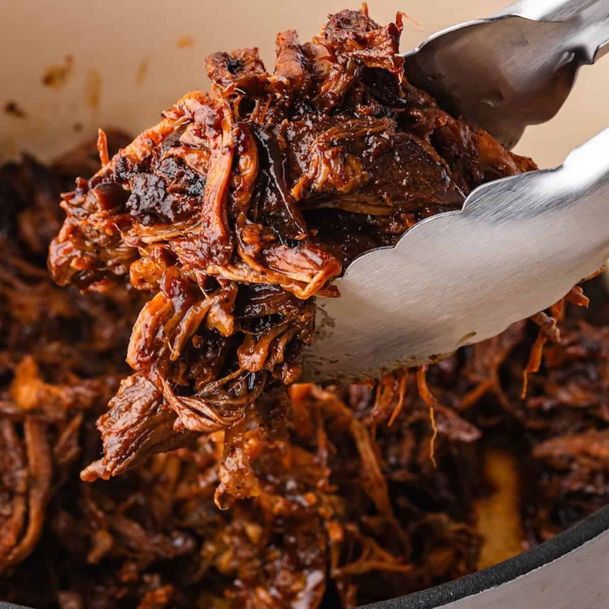Tongs lift tender, juicy shredded barbacoa beef from a Dutch oven, revealing its rich color and texture.