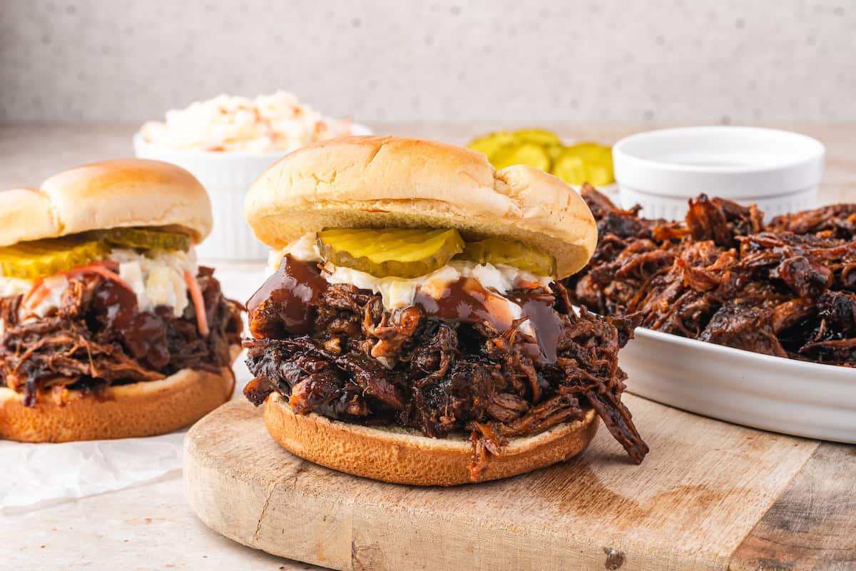 Pulled pork sandwiches with pickles and coleslaw rest on a wooden board, complemented by bowls of extra pulled pork, all slow-cooked to perfection in a Dutch oven.