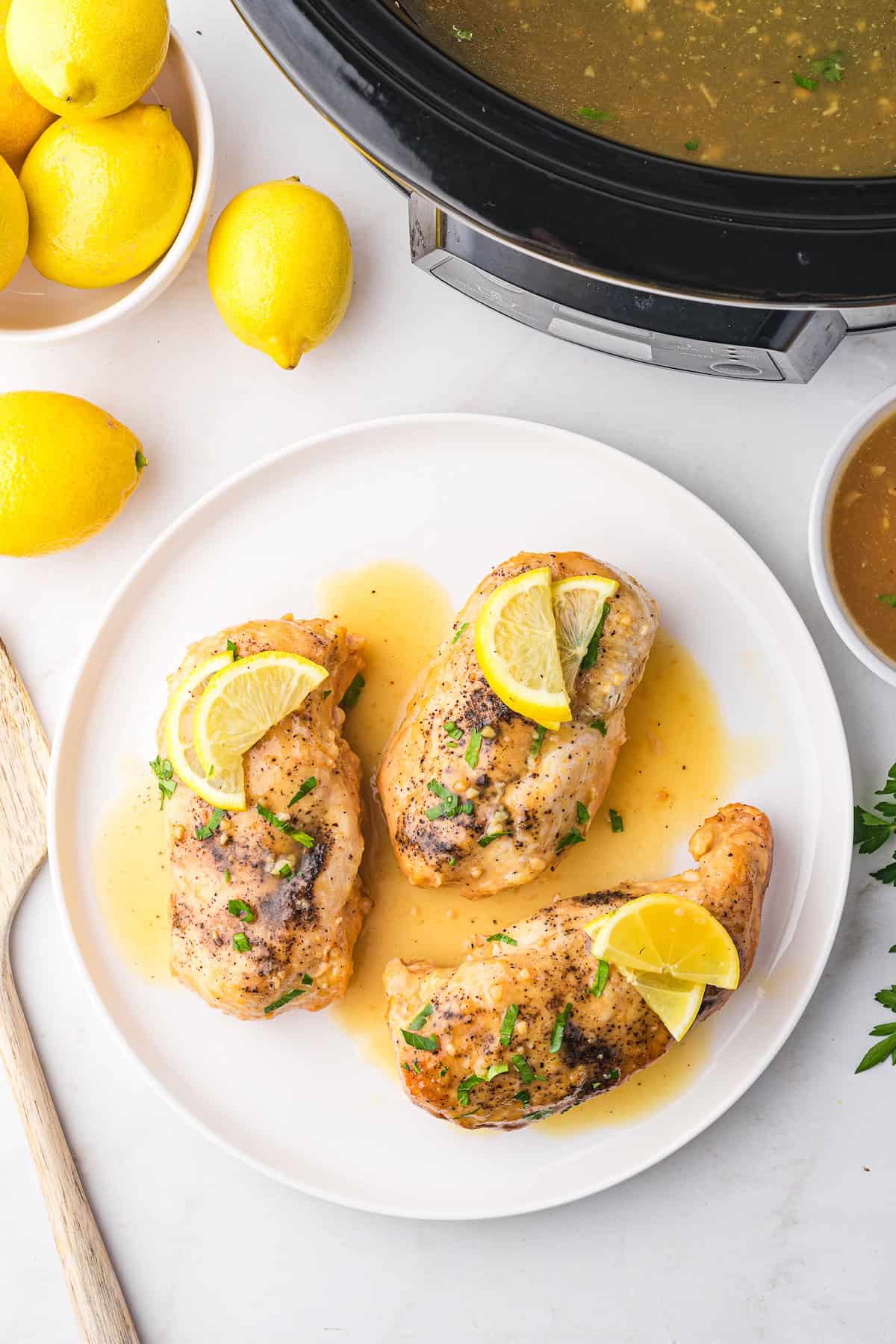 Three lemonade chicken breasts, garnished with lemon slices, rest on a white plate beside a wooden spoon and fresh lemons. A slow cooker filled with savory broth completes the scene.